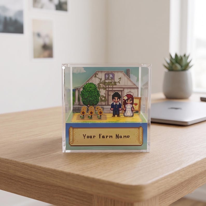 May include: A pixelated diorama in a clear acrylic cube. It depicts a wedding scene with a house, tree, and sunflowers. The text "Your Farm Name" is displayed on a banner at the bottom. The scene is set on a wooden desk.