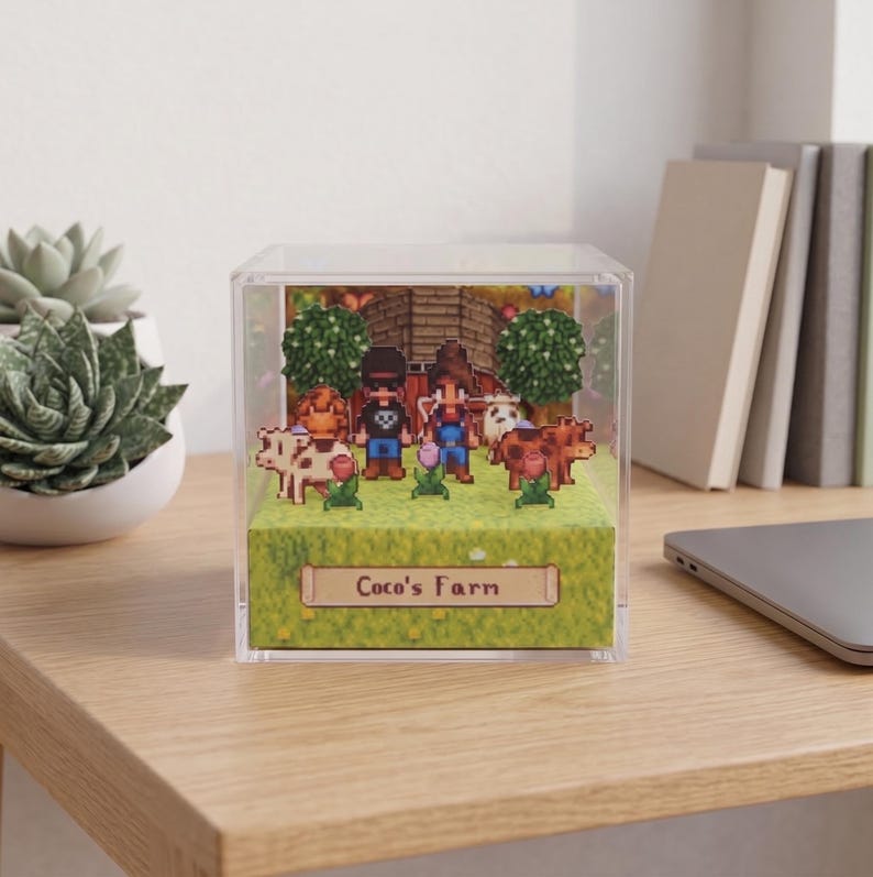 May include: A pixelated diorama inside a clear cube featuring a farm scene with two figures, cows, trees, and a barn. The diorama has the text "Coco's Farm" on a sign at the bottom. The scene is set on a green grassy base.