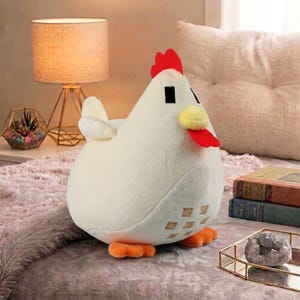 May include: A soft, white chicken toy with a red comb, yellow beak, and orange feet. The plushie has a rounded body and a pixelated design on its side. It is placed on a grey surface.