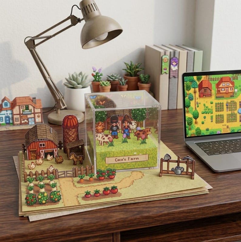 May include: A miniature farm diorama with a barn, silo, and crops, displayed on a desk with a laptop showing a farm scene. A clear acrylic box contains a pixelated farm scene with cows and farmers. A desk lamp and potted plants are also visible.