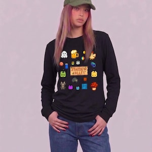以下が含まれることがあります： ピクセル化されたStardew Valleyゲームグラフィックが特徴の黒い長袖シャツ。シャツには、さまざまなゲームアイコンと「Stardew Valley」のテキストが表示されています。緑色のキャップとブルージーンズを着用しています。