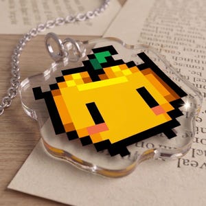 May include: A clear acrylic pendant with a pixelated yellow pumpkin design. The pumpkin has black outlines, two black rectangles for eyes, and a green stem. It hangs from a silver chain.