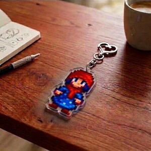 May include: A pixelated keychain featuring a character with a red hat and scarf, and a blue outfit. The keychain is attached to a silver heart-shaped clasp. The background includes a wooden surface, a notebook, a pen, and a coffee cup.