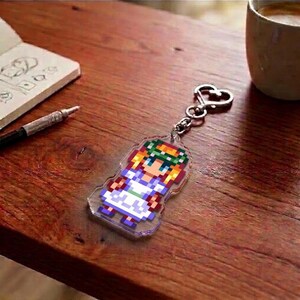 May include: A clear acrylic keychain featuring a pixelated character design. The character has blonde hair, a green wreath, and a white and blue dress. The keychain is attached to a silver-colored heart-shaped clasp.