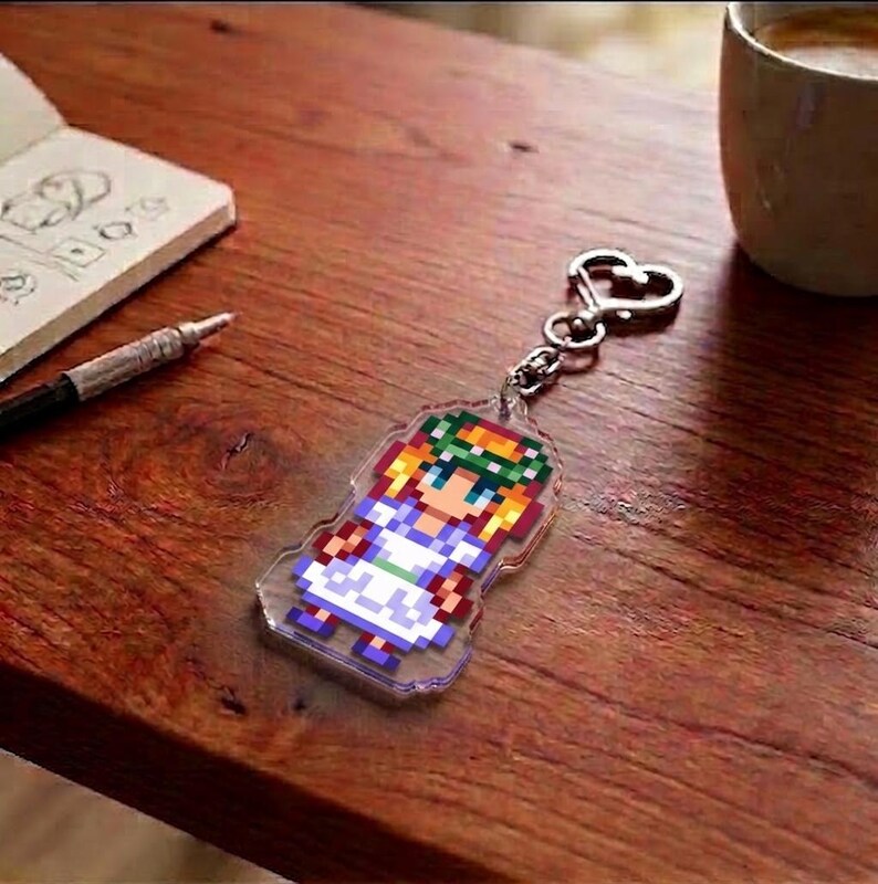 May include: A clear acrylic keychain featuring a pixelated character design. The character has blonde hair, a green wreath, and a white and blue dress. The keychain is attached to a silver-colored heart-shaped clasp.