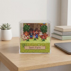 May include: A clear acrylic display cube featuring a pixelated farm scene. The scene includes figures, cows, trees, and a barn, with the text "Coco's Farm" on a sign. The cube sits on a wooden surface.