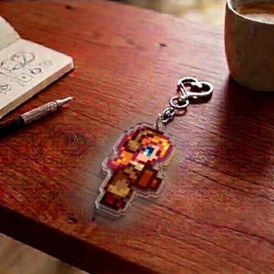 May include: A pixelated keychain featuring a character with orange hair, blue eyes, and a brown outfit. The keychain is attached to a silver heart-shaped clasp. The image is on a wooden surface.