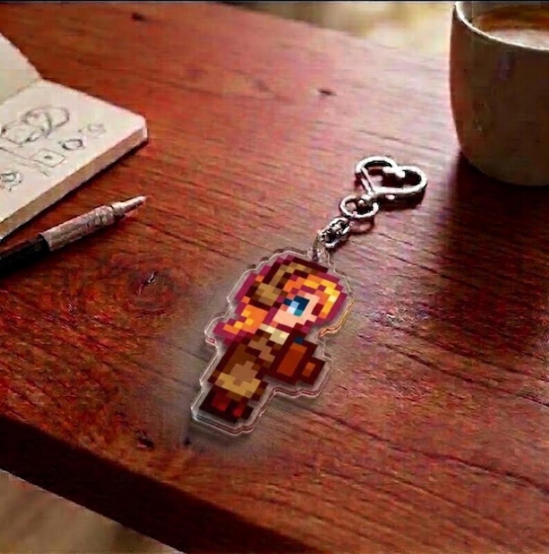 May include: A pixelated keychain featuring a character with orange hair, blue eyes, and a brown outfit. The keychain is attached to a silver heart-shaped clasp. The image is on a wooden surface.