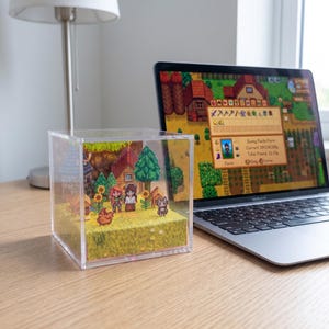 May include: A clear acrylic cube diorama featuring a pixelated farm scene with characters, houses, trees, and animals. A laptop displaying a matching game scene sits beside the cube on a wooden desk. A lamp is in the background.