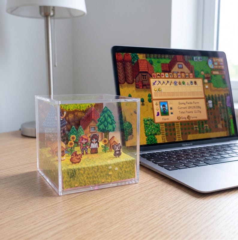 May include: A clear acrylic cube diorama featuring a pixelated farm scene with characters, houses, trees, and animals. A laptop displaying a matching game scene sits beside the cube on a wooden desk. A lamp is in the background.