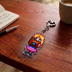 May include: A pixelated keychain featuring a character with brown hair, a blue shirt, and a pink skirt. The keychain has a silver heart-shaped clasp. The image is on a wooden surface with a notebook, pen, and a coffee cup.
