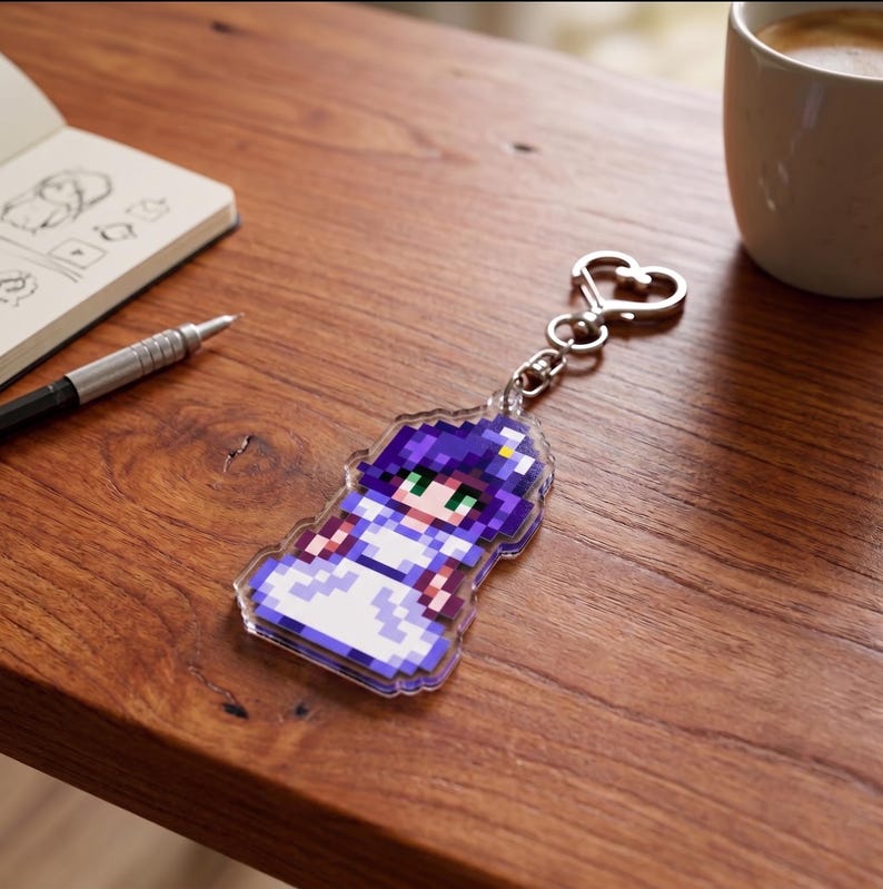 May include: A clear acrylic keychain featuring a pixelated image of a character in a white dress with purple hair and green eyes. The keychain has a silver-colored heart-shaped clasp. The background includes a notebook, pen, and a white mug on a wooden surface.