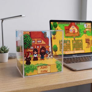 May include: A pixel art diorama featuring a farm scene with characters, animals, and a house. The diorama is in a clear acrylic box with the words "JUNO FARM" on a sign. A laptop displaying a farm game is in the background.