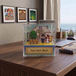 May include: A clear acrylic display case featuring a pixelated farm scene with a house, tree, and couple. The base has a banner that reads "Your Farm Name". Three framed pixel art images hang on the wall behind the case.