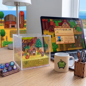 May include: A clear acrylic cube diorama featuring a pixelated farm scene with characters, trees, and a chicken. A laptop displaying a similar game scene, a mug with a pixelated apple, and a wooden pen holder are also on the desk.