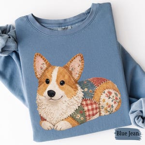 May include: A blue jean-colored sweatshirt featuring a detailed appliqué of a corgi dog. The corgi has brown and white fur, with a patchwork body of floral and plaid patterns. The word "COMFORT" is printed on the collar.