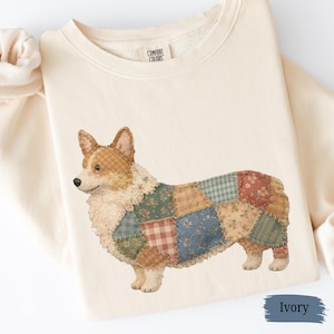 May include: Ivory-colored sweatshirt featuring a detailed illustration of a corgi with a patchwork design. The corgi's body is composed of various fabric patterns in shades of blue, green, red, and tan. The sweatshirt has a crew neck and the tag reads "Comfort Colors."