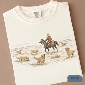 May include: Ivory-colored t-shirt featuring a detailed illustration of a cowboy riding a horse, surrounded by several golden retriever dogs. The shirt has a classic crew neck and a tag that reads "Comfort Colors".