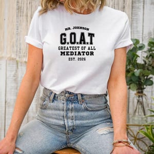May include: White t-shirt featuring the text "MR. JOHNSON G.O.A.T GREATEST OF ALL MEDIATOR EST. 2026" in black. The shirt is worn with blue jeans.