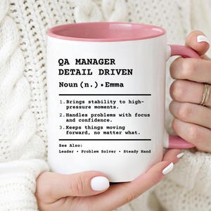May include: White ceramic mug with a pink interior and handle. The mug features the text "QA MANAGER DETAIL DRIVEN" and a definition, including "Brings stability to high-pressure moments." A perfect gift for a quality assurance professional.