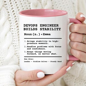 May include: White ceramic mug with a pink interior and handle. The mug features the text "DEVOPS ENGINEER BUILDS STABILITY" and a definition, including "Noun (n.) - Emma." The mug is held by a person.