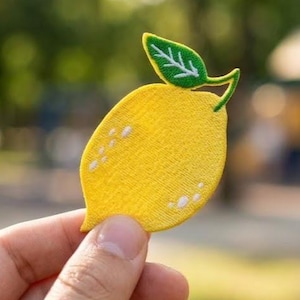 May include: A yellow lemon-shaped embroidered patch featuring a green leaf at the top. The lemon has a textured surface with small white stitched details on the side, creating a distinct fruit shape with a stem and a single leaf attached.
