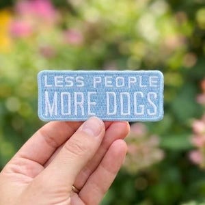 May include: A rectangular embroidered patch in light blue with white stitched text that reads LESS PEOPLE MORE DOGS. The patch features a clean, horizontal design with a border and clear, block-style lettering centred on the fabric surface.