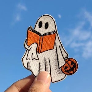 May include: An embroidered patch featuring a white ghost with black eyes holding an open orange book in one hand and a small orange jack-o'-lantern with a carved face in the other hand, outlined in black thread against a white fabric base.