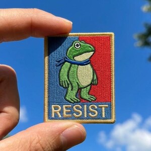 Può includere: Toppa ricamata con una rana verde con una sciarpa blu, su sfondo rosso e blu. La parola "RESIST" è ricamata in bianco in basso. La toppa è rettangolare con un bordo dorato.