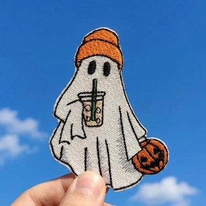 May include: An embroidered patch featuring a white ghost wearing an orange beanie. The ghost holds a clear plastic cup with a green straw containing a light brown beverage and carries a small orange jack-o'-lantern pumpkin in its other hand.