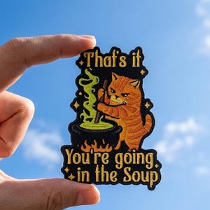 May include: An embroidered patch with a black background featuring an orange tabby cat stirring a black cauldron with green bubbling liquid. The text reads That's it You're going in the Soup in gold lettering with small gold star accents.