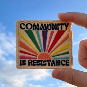 May include: A square embroidered patch with a cream-colored background featuring a rising sun design with multicolored rays in yellow, green, blue, purple, pink, and red. The black text reads COMMUNITY at the top and IS RESISTANCE at the bottom.