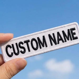 May include: A rectangular white embroidered patch with a stitched border featuring the text CUSTOM NAME in bold black capital letters. The patch has a clean, uniform fabric texture and is presented against a clear blue sky background.