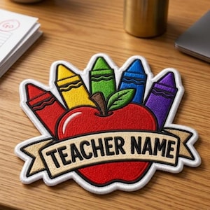 May include: An embroidered patch featuring a bright red apple with a green leaf, surrounded by five colorful crayons in red, yellow, green, blue, and purple. A beige banner across the apple displays the black text TEACHER NAME.