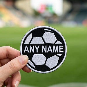 May include: A round, black and white patch featuring a soccer ball design. The text "ANY NAME" is displayed in white, centered within the ball. The patch is held against a blurred green background, suggesting a sports field.