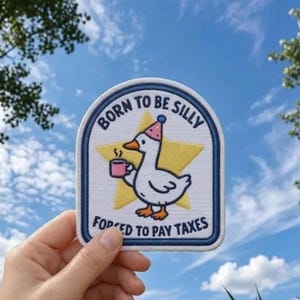 Op de afbeelding: Een witte geborduurde patch met een blauwe rand met een cartoon gans die een roze feesthoedje draagt en een roze mok vasthoudt. Achter de gans staat een gele ster. De tekst luidt BORN TO BE SILLY FORCED TO PAY TAXES in blauwe letters.