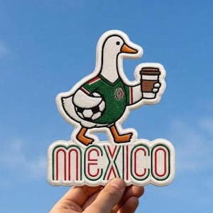 May include: An embroidered patch featuring a white duck wearing a green soccer jersey with a soccer ball under its wing and holding a brown coffee cup. Below the duck, the word MEXICO is stitched in red and green outlined letters.