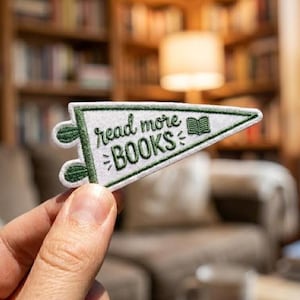 Puede incluir: Un parche blanco en forma de banderín con ribetes y detalles en verde. El parche dice "read more BOOKS" en una fuente de estilo manuscrito. Un pequeño libro bordado está en el lado derecho. El parche se sostiene en una mano.