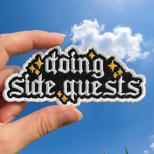 May include: An embroidered patch with a black background and white text that reads "doing side quests" in a gothic-style font. The patch features small yellow four-pointed star accents and a thick white border around the entire shape.