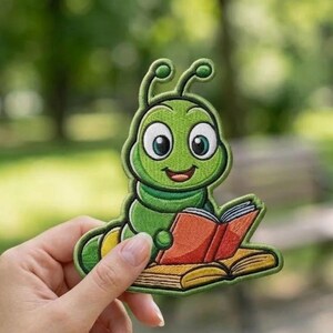 May include: An embroidered patch featuring a bright green cartoon caterpillar with large white eyes and antennae, sitting and reading an open book with orange-red covers and yellow pages, finished with a dark green stitched border.