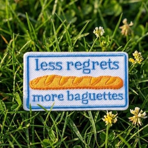 Può includere: Una toppa rettangolare ricamata con sfondo bianco e bordo blu. Il testo "less regrets" è scritto in blu sopra un'illustrazione di una baguette arancione, con il testo "more baguettes" in blu sotto il disegno del pane.