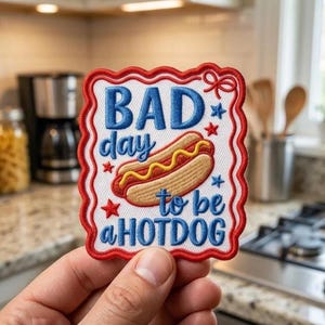 May include: Embroidered patch with a red, white, and blue design. The patch features the text "BAD day to be a HOTDOG" with a cartoon hot dog illustration. The patch has a red scalloped border and star accents.