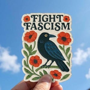 Puede incluir: Un parche bordado rectangular con fondo color crema que muestra un cuervo negro posado sobre un tallo verde. El parche presenta el texto FIGHT FASCISM en letras mayúsculas negras, rodeado de vibrantes flores de amapola rojo anaranjado.