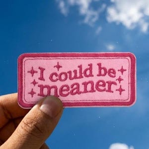 May include: A rectangular pink embroidered patch with a dark pink border. The text "I could be meaner" is stitched in dark pink in the center, surrounded by small four-pointed star icons on the left and right sides of the lettering.