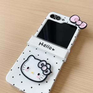 Op de afbeelding: Witte telefoonhoes met zwarte stippen, met een Hello Kitty-ontwerp. De hoes heeft een roze strikaccent en het woord "Hello" erop gedrukt. Het telefoonscherm is zwart.