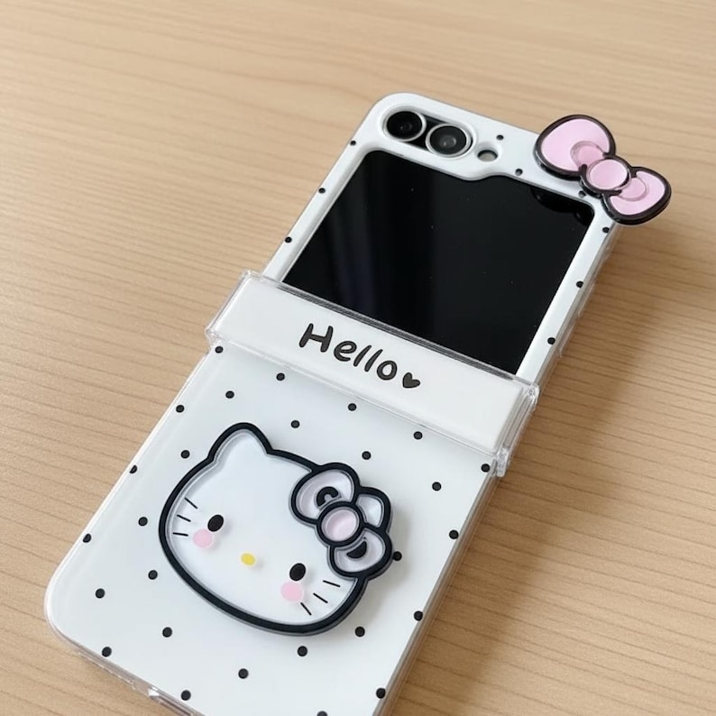 Op de afbeelding: Witte telefoonhoes met zwarte stippen, met een Hello Kitty-ontwerp. De hoes heeft een roze strikaccent en het woord "Hello" erop gedrukt. Het telefoonscherm is zwart.