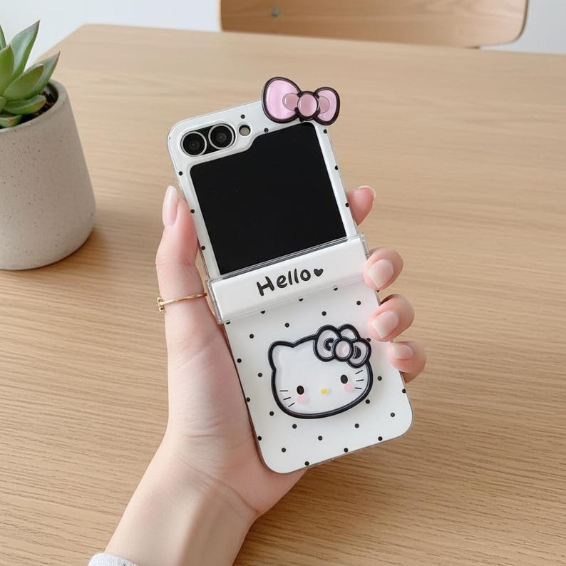 Op de afbeelding: Witte telefoonhoes met een Hello Kitty-ontwerp. De hoes heeft een zwart-wit stippenpatroon, een Hello Kitty-gezicht en een roze strik. De telefoon is open en toont een zwart scherm. Het woord "Hello" staat op de hoes.