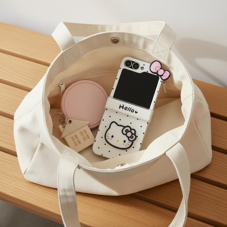 Op de afbeelding: Een cr&egrave;mekleurige canvas draagtas met ritssluiting, met een Hello Kitty telefoonhoesje, een roze compacte spiegel en een sleutelhanger in de vorm van een huis. De telefoonhoes is wit met zwarte stippen en een Hello Kitty-ontwerp. Op de telefoonhoes staat "Hello".