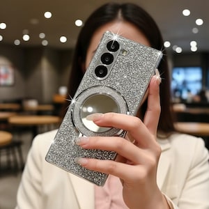 May include: A sparkly silver phone case with a three-lens camera cutout and a circular ring holder. The case is covered in glitter and has a reflective mirror in the center of the ring. The phone case is rectangular and has rounded corners.
