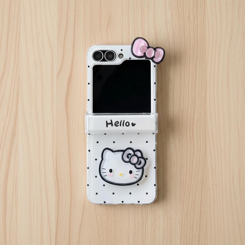Op de afbeelding: Witte telefoonhoes met een Hello Kitty-ontwerp. De hoes heeft een zwart scherm, een roze strik en een Hello Kitty-gezicht met een strik. De hoes is versierd met zwarte stippen.
